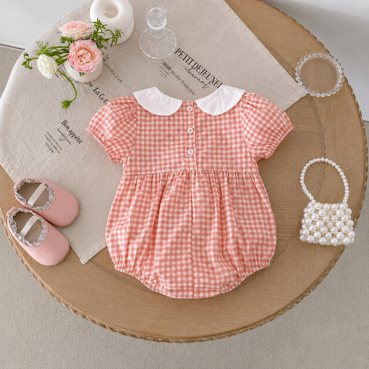 Pink gingham baby romper with white collar on a wooden surface, surrounded by baby shoes, flowers, and a small handbag.