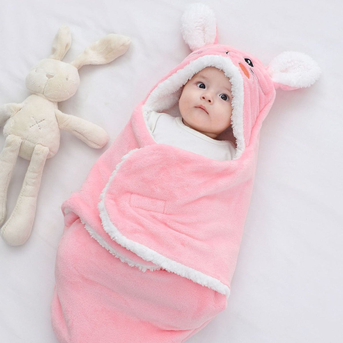 Baby wrapped in a pink swaddle with bunny ears, lying next to a plush toy on a light background