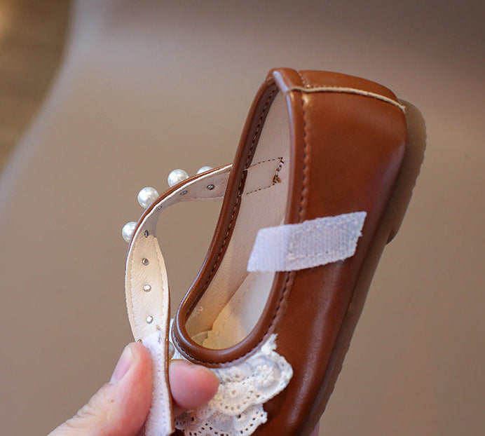 Brown shoe with white lace and pearls held by a hand against a neutral background