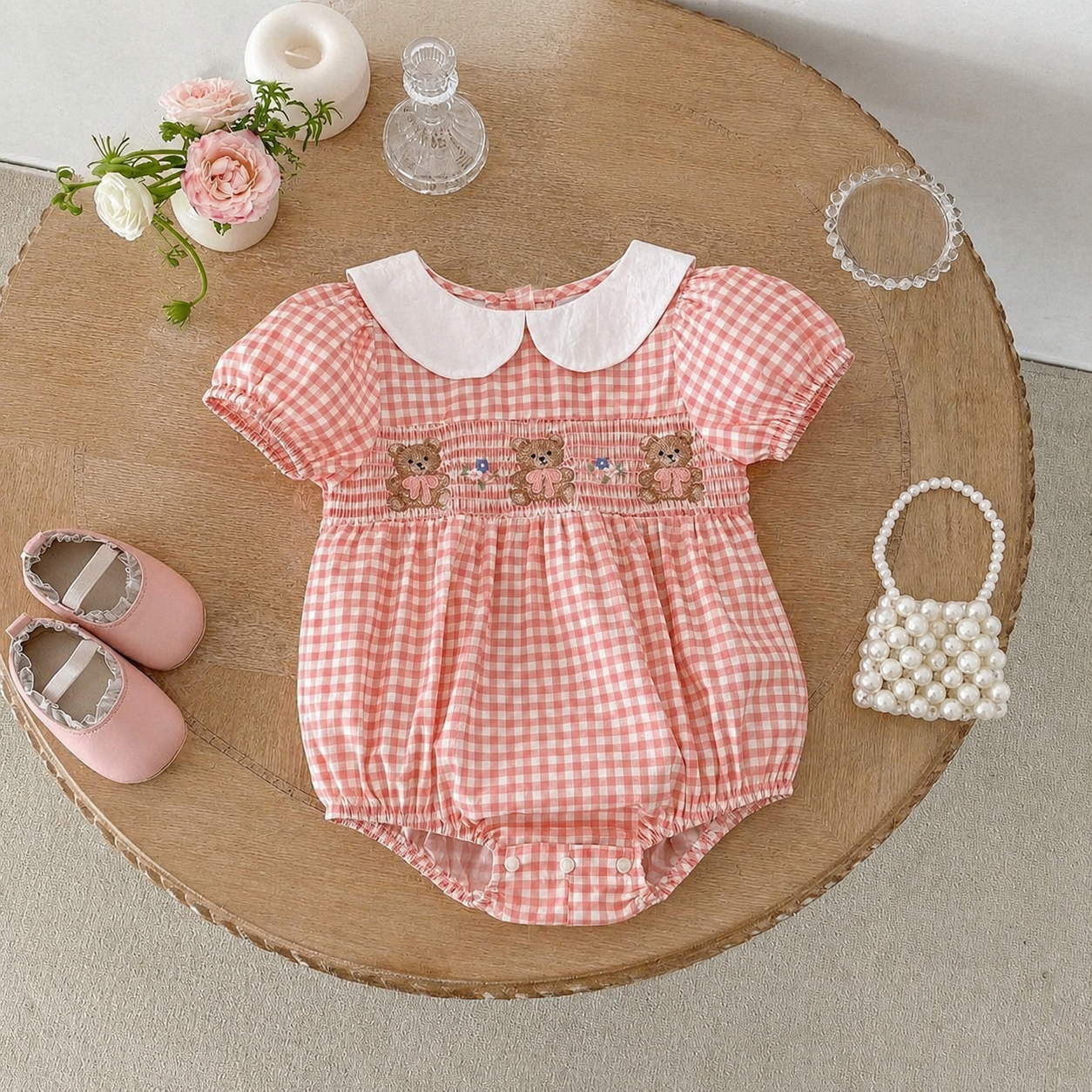 Pink checkered baby romper with teddy bear design on a wooden table with pink shoes and a small bag.