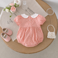 Pink gingham baby romper with white collar on a wooden surface, surrounded by baby shoes, flowers, and a small handbag.