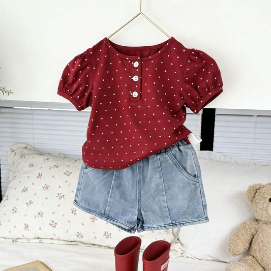 Red polka dot top, denim shorts, and red boots on a bed with a teddy bear and book.