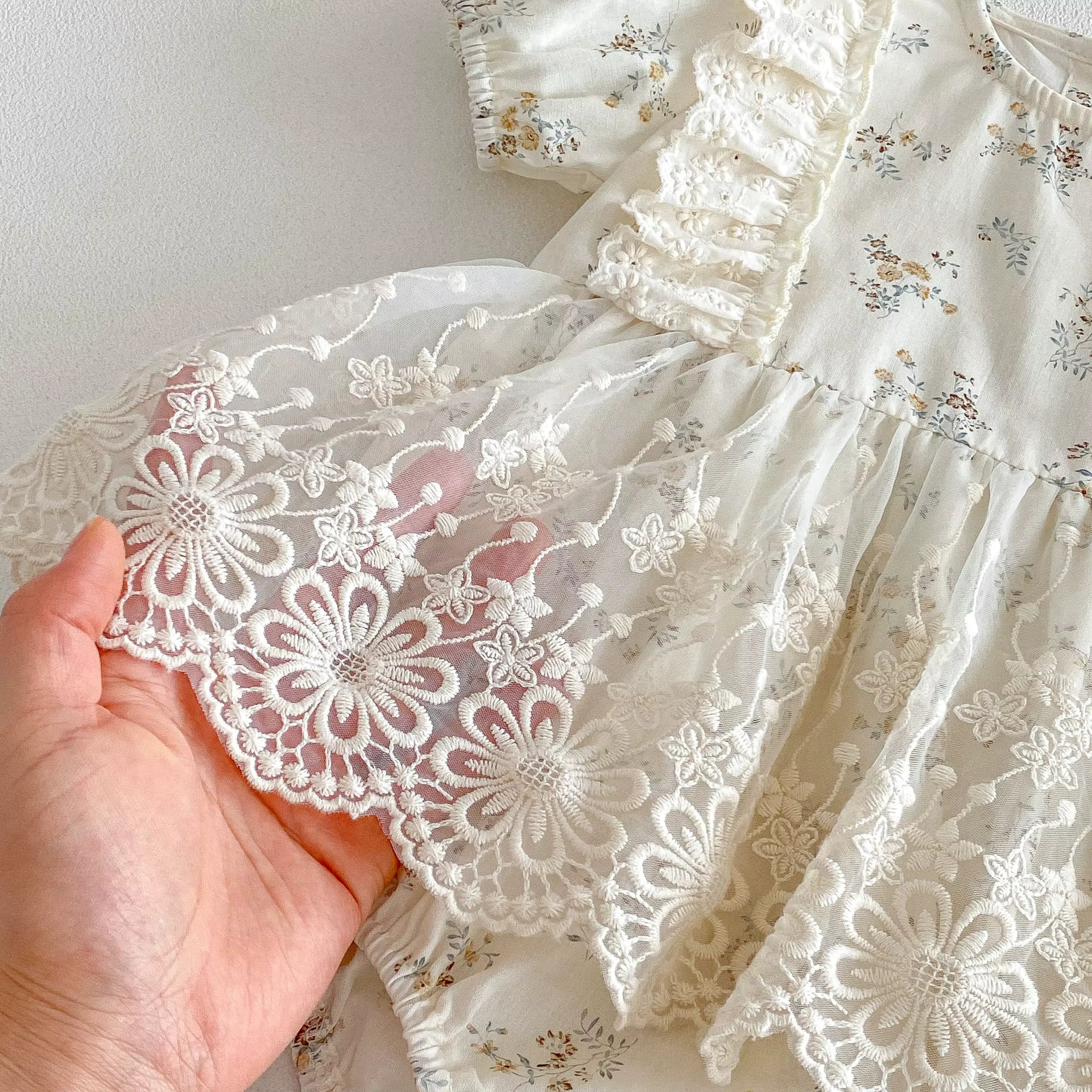 Close-up of a hand holding a piece of lace fabric with a floral dress in the background.