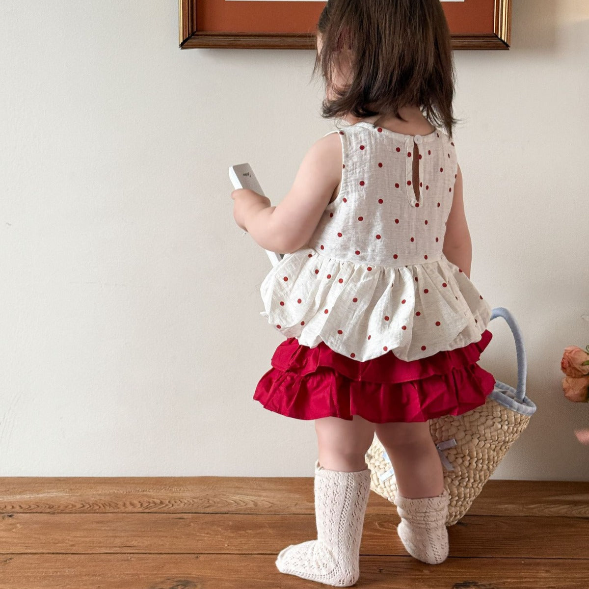 Child wearing a white top with red polka dots and a red skirt, standing on a wooden floor.