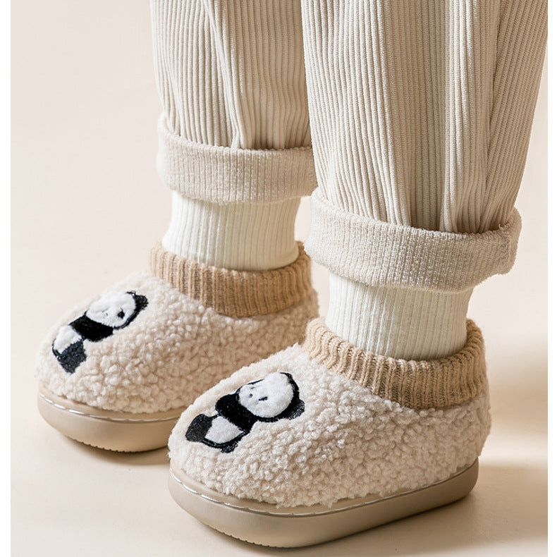 White slippers with panda design worn by a person in white socks and beige pants.