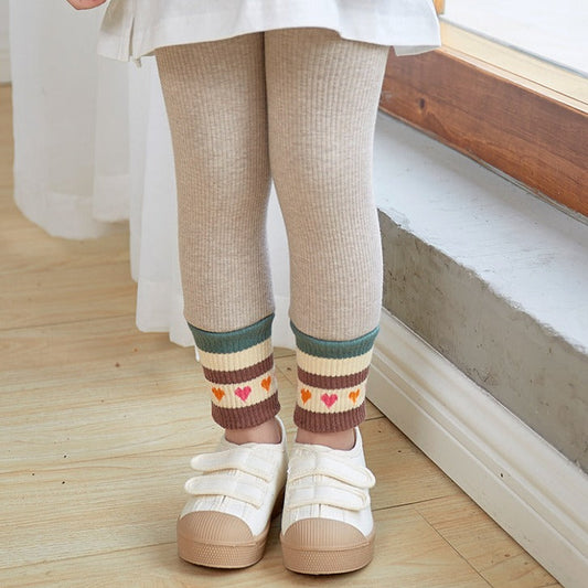 Child wearing beige leggings with colorful heart patterns and white shoes indoors.