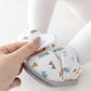 A close-up of a baby's foot wearing a white soft bootie with a colorful geometric and animal print, while an adult hand adjusts the strap.