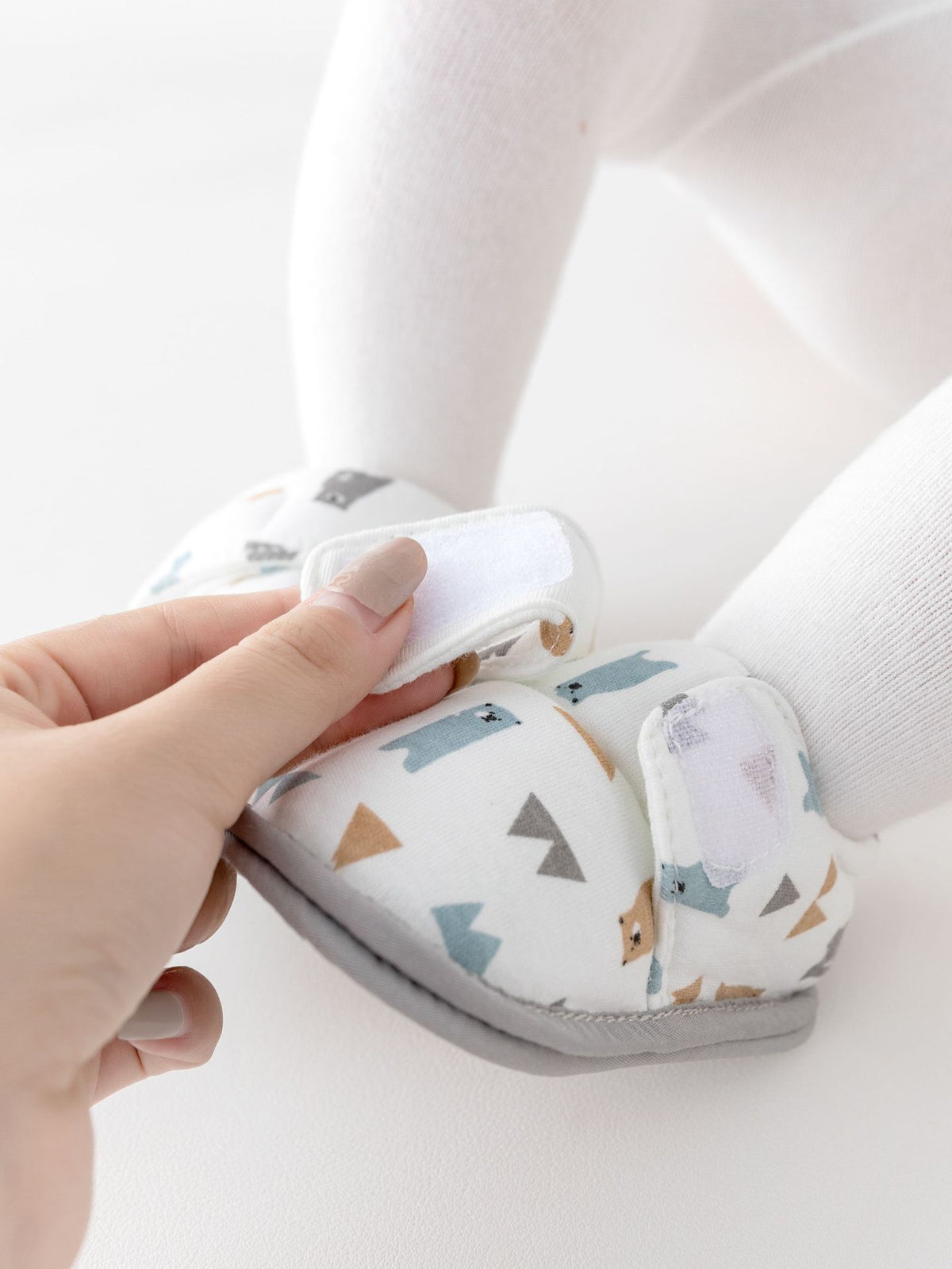 A close-up of a baby's foot wearing a white soft bootie with a colorful geometric and animal print, while an adult hand adjusts the strap.