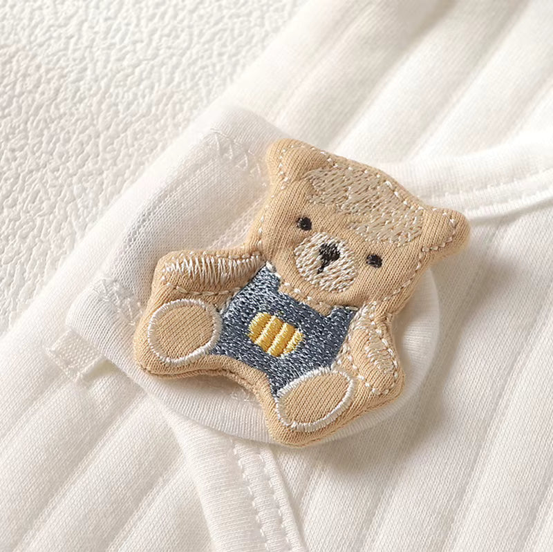 A close-up of a white ribbed fabric garment featuring an embroidered teddy bear applique. The bear is beige with dark brown embroidered eyes and nose, wearing a blue denim-style overalls outfit with yellow striped details.